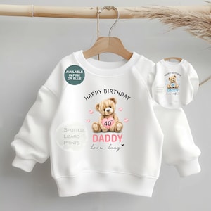 May include: White sweatshirt with a teddy bear graphic holding a pink heart with the number 40. The text reads "Happy Birthday Daddy Love Lucy". A smaller version of the sweatshirt is also visible. The text "Available in Pink or Blue" is also present.