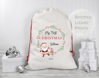 May include: A white Christmas stocking with a red drawstring. The stocking features a green wreath with red berries and a cartoon Santa Claus. The text "My First Christmas William 2021" is printed on the stocking.