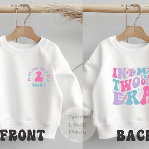 Könnte beinhalten: Weißes Sweatshirt mit einem Design für den Geburtstag eines Zweijährigen. Auf der Vorderseite steht "In my birthday era" mit der Zahl 2 und einem Namen. Auf der Rückseite steht "In my two year old era" in Retro-Schrift.