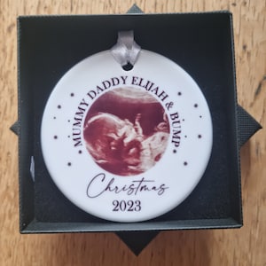 Może przedstawiać: Okrągła biała ceramiczna ozdoba ze srebrną wstążką, z centralnie umieszczonym zdjęciem USG. Tekst głosi MUMMY DADDY ELIJAH & BUMP, Christmas oraz 2023, w otoczeniu małych gwiazdek. Ozdoba jest zaprezentowana w czarnym pudełku.