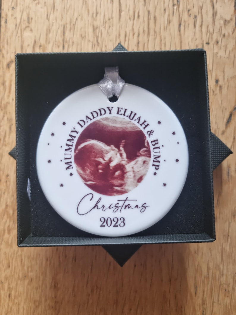 Może przedstawiać: Okrągła biała ceramiczna ozdoba ze srebrną wstążką, z centralnie umieszczonym zdjęciem USG. Tekst głosi MUMMY DADDY ELIJAH & BUMP, Christmas oraz 2023, w otoczeniu małych gwiazdek. Ozdoba jest zaprezentowana w czarnym pudełku.