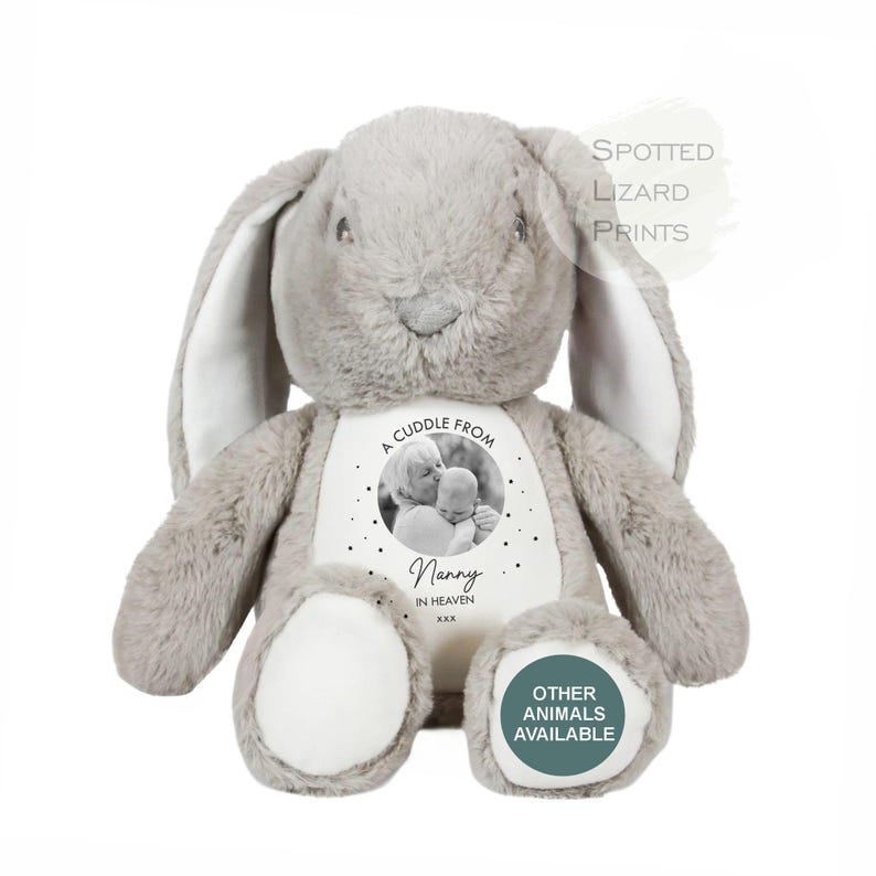 May include: A plush grey rabbit toy with long ears and white foot pads. The chest features a circular black and white photo of a person holding a baby, with the text A CUDDLE FROM Nanny IN HEAVEN xxx. The text SPOTTED LIZARD PRINTS appears above.