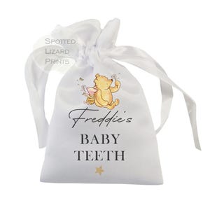 May include: White satin drawstring bag with Winnie the Pooh and Piglet characters. The bag has the text "Freddie's Baby Teeth" printed on it.