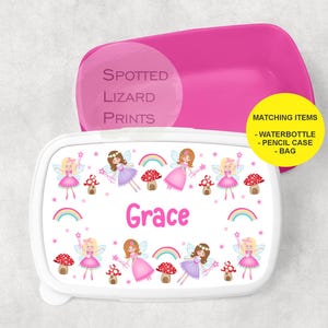 Könnte beinhalten: Weiße Lunchbox mit rosa Deckel. Die Lunchbox hat ein Design mit Feen mit Regenbogen-Akzenten und dem Namen "Grace" auf der Vorderseite. Der Deckel trägt den Text "Spotted Lizard Prints" und einen gelben Aufkleber mit der Aufschrift "Passende Artikel - Wasserflasche - Stifteetui - Tasche".