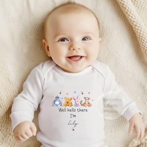 Op de afbeelding: Een witte babybody met lange mouwen. Het heeft een kleurrijke illustratie van Winnie de Poeh-personages. De tekst "Well hello there I'm Lily" staat eronder gedrukt, met een bij.