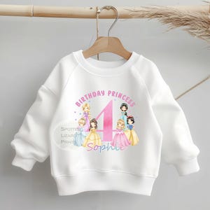 Könnte beinhalten: Weißes Sweatshirt mit einem rosa-lila Aufdruck mit der Aufschrift "Birthday Princess 4 Sophie". Die Grafik zeigt Illustrationen mehrerer Prinzessinnen. Das Sweatshirt hängt an einem Holzkleiderbügel.