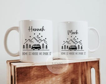 May include: Two white ceramic mugs with black graphic designs. One mug says "Hannah" and the other says "Mark". Both mugs feature a mountain, trees, a camper van, and the text "HOME IS WHERE WE PARK IT".