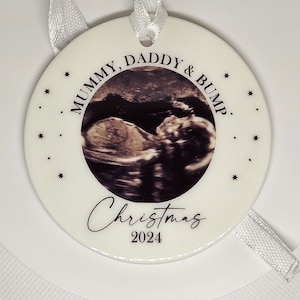 Może przedstawiać: Okrągła biała ceramiczna ozdoba z centralnie umieszczonym zdjęciem USG. Tekst głosi Mummy, Daddy & Bump, Christmas oraz 2024. Wok&oacute;ł obrazka nadrukowano małe czarne gwiazdki. Do zawieszenia służy dołączona biała wstążka.