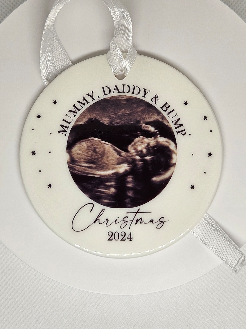 Może przedstawiać: Okrągła biała ceramiczna ozdoba z centralnie umieszczonym zdjęciem USG. Tekst głosi Mummy, Daddy & Bump, Christmas oraz 2024. Wok&oacute;ł obrazka nadrukowano małe czarne gwiazdki. Do zawieszenia służy dołączona biała wstążka.