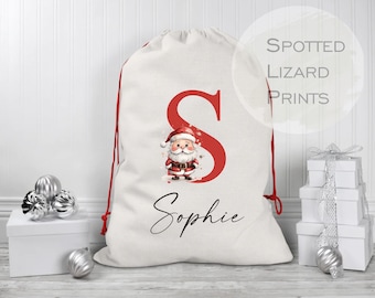 Puede incluir: Un saco de Santa de lona blanca con cordones de extracción rojos. El saco presenta una letra roja "S" con un Papá Noel de dibujos animados en su interior. El nombre "Sophie" está impreso en cursiva negra debajo de la letra. El texto "Spotted Lizard Prints" es visible en un círculo blanco en la esquina superior derecha de la imagen.