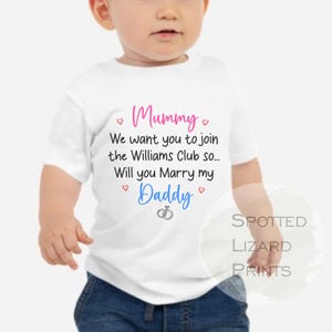 May include: A white short-sleeved baby t-shirt featuring the pink text Mummy, black text We want you to join the Williams Club so... Will you Marry my, blue text Daddy, and small red hearts and a black line drawing of two interlocking wedding rings.