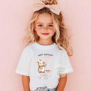 May include: A white t-shirt with the text "HAPPY BIRTHDAY" and a graphic of a cartoon animal holding a balloon with the number 7. The shirt also has the name "Hattie" and a small cupcake graphic. The shirt is from Spotted Lizard Prints.