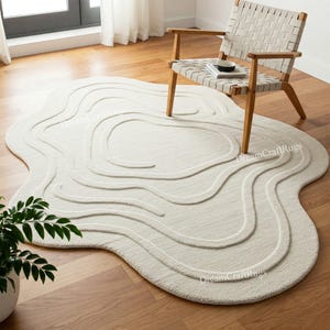 Op de afbeelding: Een wit, pluche, abstract vloerkleed met een golvend, organisch ontwerp. Het vloerkleed ligt op een houten vloer met een houten stoel op de achtergrond.