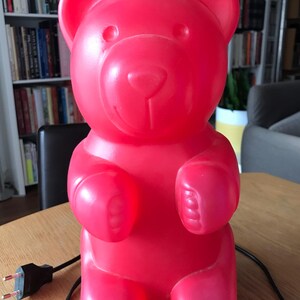 Op de afbeelding: Een felrode gummybeer-lamp. De lamp heeft de vorm van een beer en heeft een glad, glanzend oppervlak. Het heeft een lachend gezicht, afgeronde oren en korte ledematen. De lamp is via een zwarte kabel aangesloten op een stopcontact.