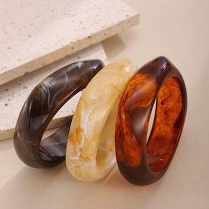 May include: Three chunky, resin bangles in shades of brown, cream, and amber. The bangles have a marbled effect, with swirls of color creating a unique pattern. The bracelets are stacked together, showcasing their varying hues and textures.