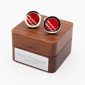 May include: A pair of round cufflinks with a red center and a white stitched design, set in a silver frame. The cufflinks rest on a wooden box with a silver nameplate that reads "Mark Robinson Est. 1964."
