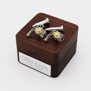 May include: A pair of silver and gold golf club cufflinks with a golf ball design, presented on a dark wooden box. The box has a silver nameplate that reads "Ollie Gale Est. 2003".