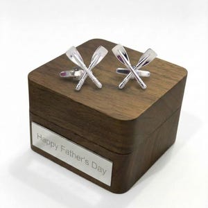 May include: Silver cufflinks in the shape of crossed oars rest on a dark wooden box. The box has a silver plate with the text "Happy Father's Day". The cufflinks are detailed with textured handles and paddle blades.