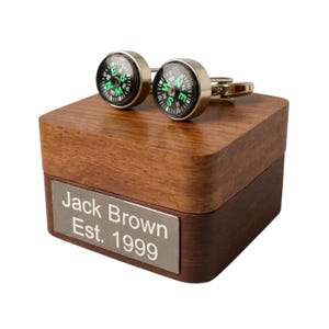 May include: Silver-toned compass cufflinks with black faces and green lettering, displayed on a two-tone wooden box. The box features a silver nameplate with the inscription "Jack Brown Est. 1999".