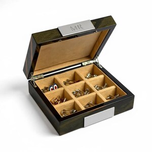Può includere: Scatola di legno aperta con finitura verde scuro e marrone, foderata in tessuto beige. La scatola è divisa in scomparti, riempiti con vari gemelli. Una placca d'argento sul coperchio è incisa con "MR".
