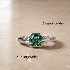 May include: A silver ring with a square, emerald-cut green gemstone. The ring has a modern design with a split shank. The text "Alcazerjewelry" is visible in the image.