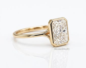 May include: A gold ring featuring a rectangular radiant-cut center stone set in a bezel setting. The band is thin and polished gold. The text AlcazerJewelry is visible in the lower right corner of the image.