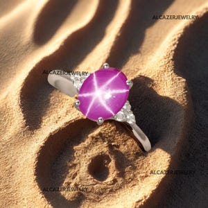 May include: A silver ring with a large, oval star ruby. The ruby is a bright pink with a six-pointed star design. Small, clear gemstones are set on the band. The ring is presented on a sandy surface.