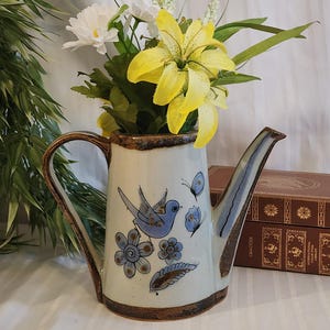 May include: A ceramic pitcher with a cream-colored glaze, featuring a hand-painted blue bird, blue flowers, and butterflies. The pitcher has a dark brown rim and base, a curved handle, and a spout, filled with artificial yellow lilies and white flowers.
