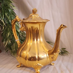 May include: A polished gold-tone metal teapot featuring a bulbous, vertically ribbed body, a curved handle with ornate detailing, a long spout, and a lid with a finial. The teapot stands on three decorative feet with shell-like motifs.