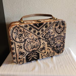 May include: Vintage rectangular suitcase with a brown leather handle and trim. The suitcase features a beige and black floral pattern on a textured fabric. The suitcase is sitting on a white surface.