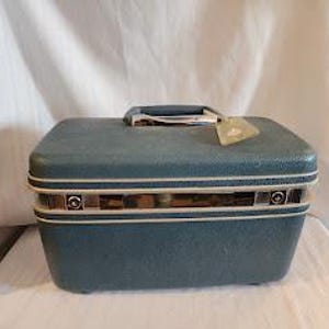 May include: A rectangular blue train case with a textured exterior, featuring a top handle, a central silver-toned metal band with two circular latches, and a light-colored trim. A small, light-colored rectangular tag is attached to the handle.