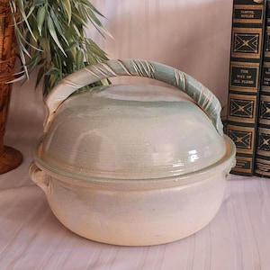 May include: A light blue and cream-colored ceramic casserole dish with a lid and a twisted handle. The dish has a round shape and small handles on either side. The lid is slightly domed. The dish is likely used for cooking and serving.
