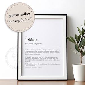 Op de afbeelding: Een zwart omkaderde print met het woord "lekker" en de definitie ervan in zwarte tekst. Een beige ronde sticker met de woorden "personalise example text" staat linksboven. Rechts staat een kleine plant in een pot.