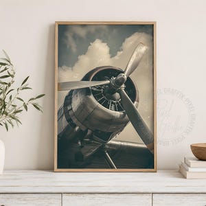 May include: A framed vertical wall art print featuring a close-up view of a vintage airplane engine with a large three-blade propeller. The metallic engine is set against a cloudy sky background, captured in a muted, desaturated color palette.