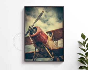 May include: A framed wall art print featuring a low-angle view of a vintage biplane with a red and cream fuselage, a prominent propeller, and wings against a cloudy sky. The artwork is enclosed in a thin black frame and displays a watermark.