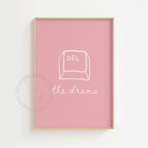 May include: A framed pink art print with a gold border. The print features a white outlined DEL key graphic above the handwritten text "the drama". The artwork is a minimalist design.