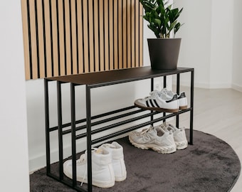 Puede incluir: Zapatero de metal negro con tres estantes, que contiene una variedad de zapatos. El zapatero está colocado sobre una alfombra ovalada gris oscuro frente a una pared con paneles de madera verticales.