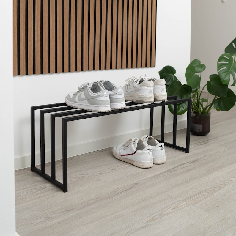 Puede incluir: Un zapatero de metal negro con dos estantes. El zapatero tiene un par de zapatillas blancas en cada estante. El zapatero est&aacute; delante de una pared con paneles de madera.