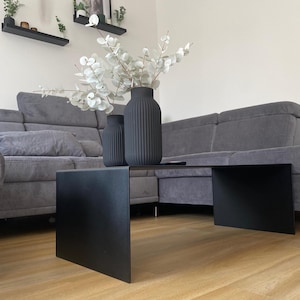 Peut inclure: Une table basse en métal noir au design unique, avec un bord surélevé d'un côté. La table est dans un salon avec un canapé gris et un vase de fleurs blanches.