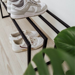 Puede incluir: Un zapatero de metal negro con tres niveles. El nivel superior tiene un par de zapatillas blancas con detalles negros. Los dos niveles inferiores tienen un par de zapatillas blancas con detalles rojos.