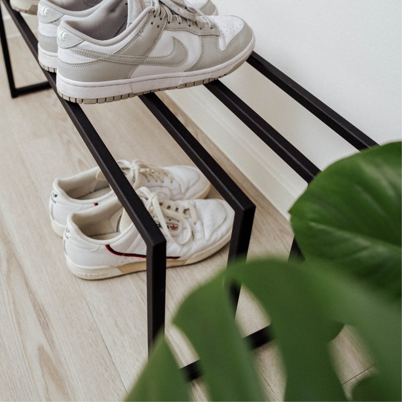 Puede incluir: Un zapatero de metal negro con tres niveles. El nivel superior tiene un par de zapatillas blancas con detalles negros. Los dos niveles inferiores tienen un par de zapatillas blancas con detalles rojos.