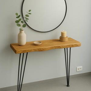 Può includere: Consolle in legno con bordo naturale e gambe a forcina nere. Uno specchio rotondo nero è appeso sopra. Il tavolo è decorato con un vaso, una piccola ciotola e una candela.