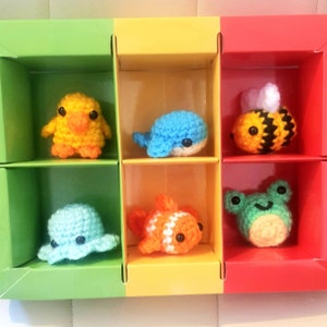 May include: Six crocheted stuffed animals in a colorful box. The animals are a yellow chick, a blue whale, a black and yellow bee, an orange and white fish, a blue octopus, and a green frog.