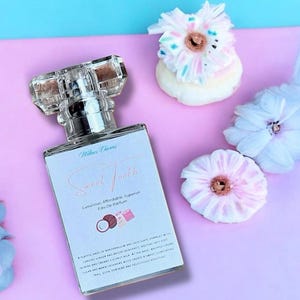 May include: A perfume bottle with a clear, geometric cap and a rectangular body. The label reads "Sweet Tooth" and includes text about the fragrance's notes. The bottle is on a pink surface, with decorative flowers.