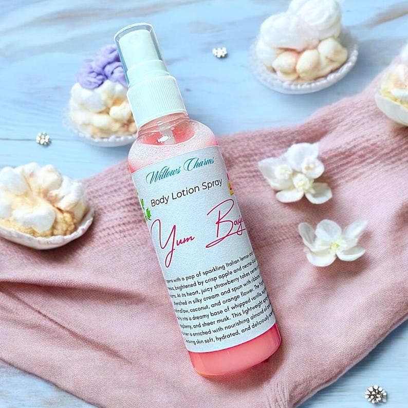May include: A pink body lotion spray bottle with the text "Willows Charms Body Lotion Spray Yum Bay" on a pink cloth. The bottle is surrounded by white flowers and meringue cookies.