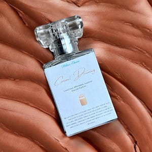 Pu&ograve; includere: Un flacone di profumo rettangolare con un tappo trasparente e sfaccettato. L'etichetta recita "Caramel Dreaming" e "Luxurious, Affordable, Superior Eau De Parfum". La bottiglia &egrave; su una superficie marrone strutturata.