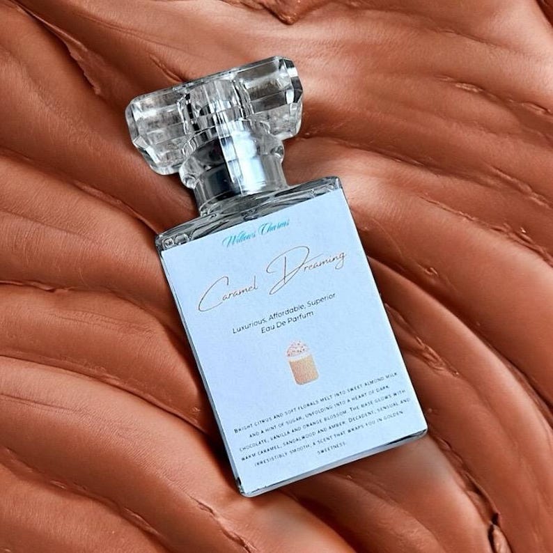 Pu&ograve; includere: Un flacone di profumo rettangolare con un tappo trasparente e sfaccettato. L'etichetta recita "Caramel Dreaming" e "Luxurious, Affordable, Superior Eau De Parfum". La bottiglia &egrave; su una superficie marrone strutturata.