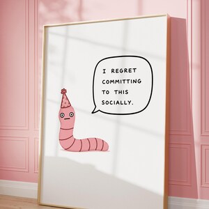 May include: A framed art print features a cartoon worm wearing a party hat and a speech bubble with the text "I REGRET COMMITTING TO THIS SOCIALLY." The worm is pink, and the background is white. The frame is gold.