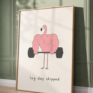 May include: A framed wall art print featuring a cartoon illustration of a pink flamingo with an extremely muscular upper body lifting a black barbell. The flamingo has thin legs and the text "leg day skipped" is printed in black below the figure.