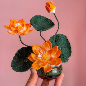 Peut inclure: Un arrangement floral au crochet fait à la main avec des fleurs de lotus orange, des feuilles vertes et un bouton, présenté dans un petit pot en céramique vert foncé. Les fleurs ont des centres blancs et jaunes. L'arrangement est sur fond rose.
